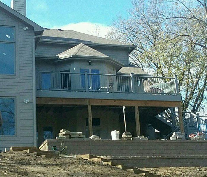 Two-story house with a wooden deck. Grey siding, blue sky, and a bare tree. Construction materials visible.