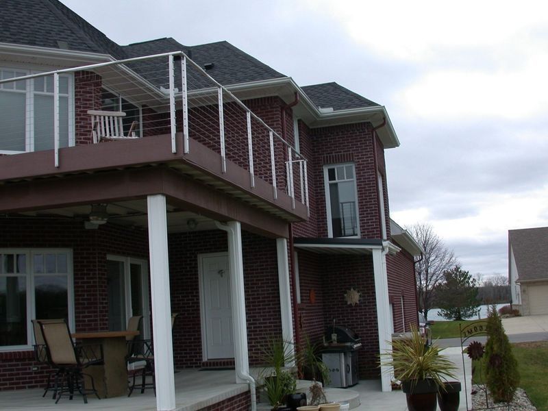 Two-story brick house with deck and metal railing, overlooking a body of water on a cloudy day.