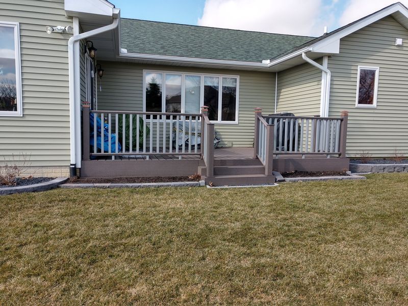 Backyard deck with gray railings, steps, and siding, overlooking a green lawn under a partly cloudy sky.