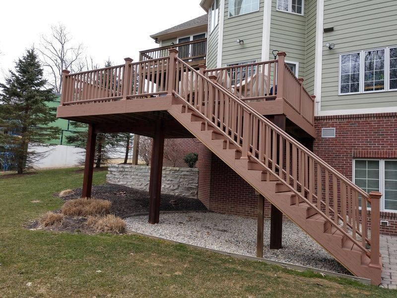 Brown deck with stairs extending from a light green house, supported by wooden posts on a grassy yard.