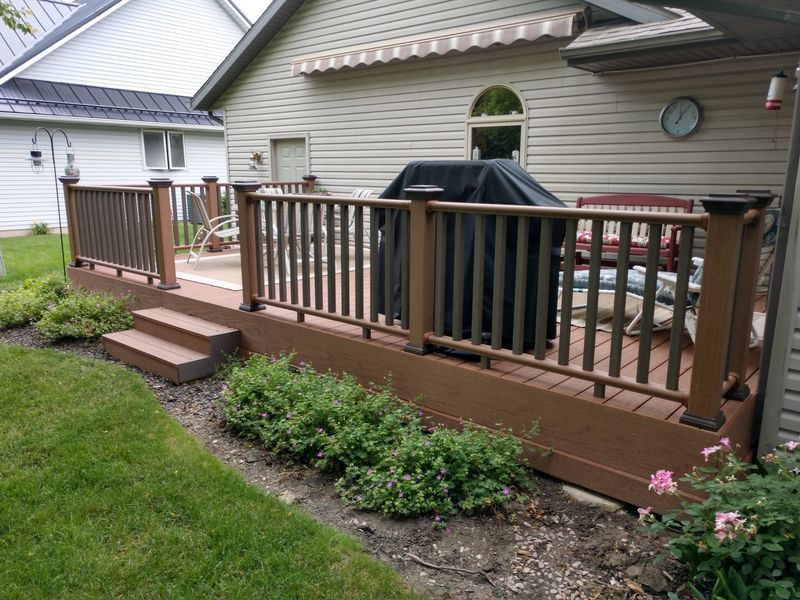 Brown wooden deck with railings, steps, and built-in grill, next to a house with beige siding and a grassy lawn.