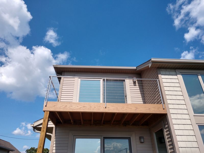 A house with a wooden deck and stainless steel railing under a blue sky with fluffy white clouds.