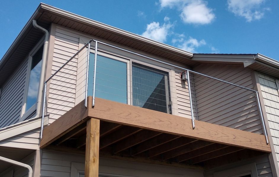 Deck with stainless steel railings on a two-story house with tan siding and blue sky.