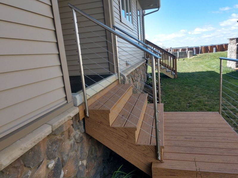 Wooden stairs and deck with metal railing next to a house with tan siding and a stone foundation.