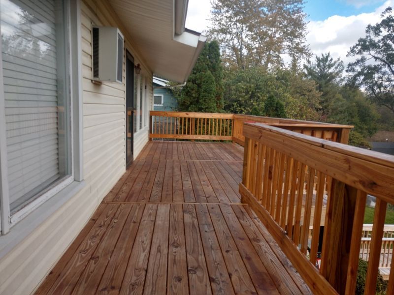 Wooden deck with railing, connected to a building with windows, overlooking trees.