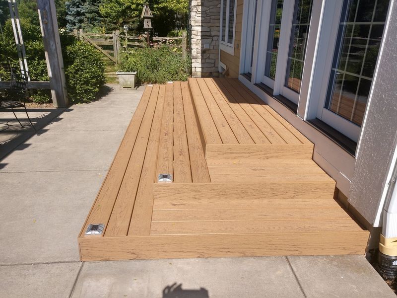 Wooden deck with steps leading to French doors, set on a concrete patio, with landscaping in the background.