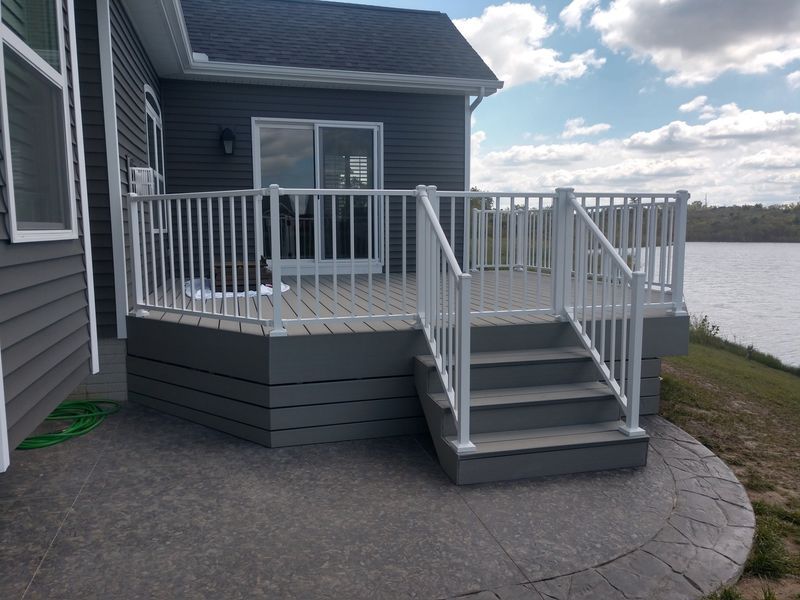 Gray deck with white railing, stairs, and a sliding glass door; next to a body of water.