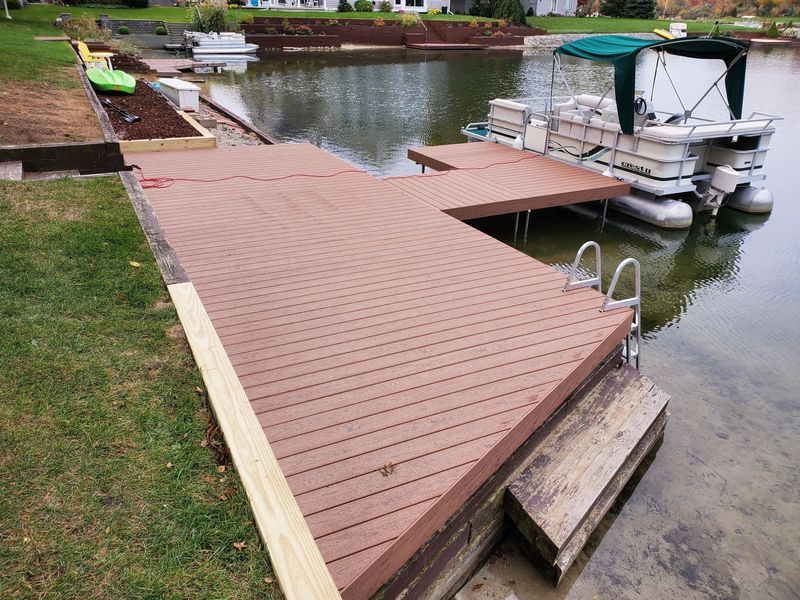 A brown composite dock extends into a lake, with a pontoon boat docked to its side.