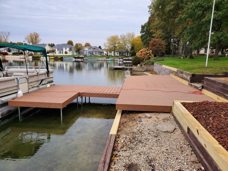 Brown composite dock extending over water, connecting to a land-based deck with raised beds; houses in the background.