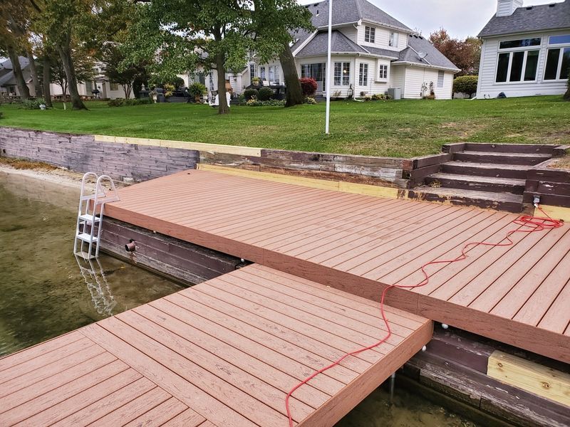 Brown composite dock on water with steps and ladder, next to a grassy yard and houses.