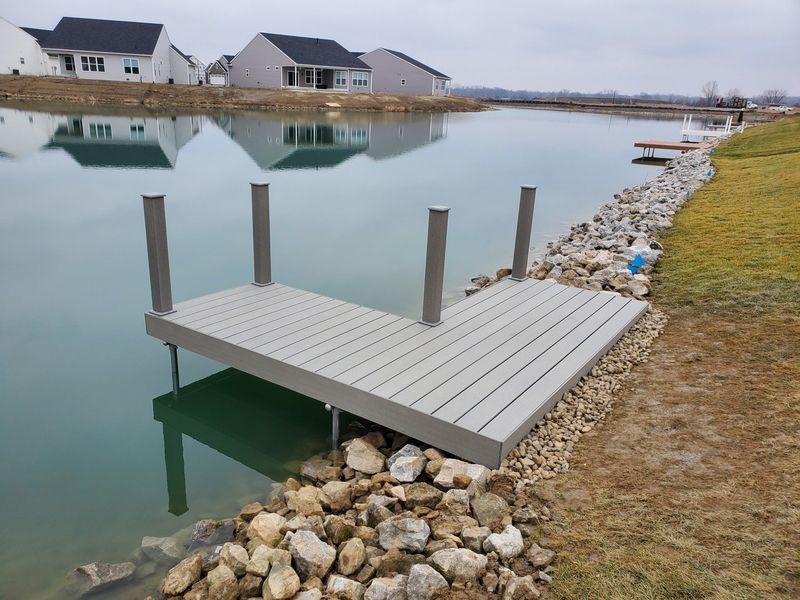 Gray dock on a pond with rocky shoreline, houses in the background.