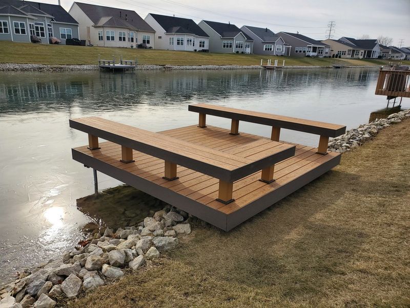 Wooden dock with seating over water, near a residential area. Brown, gray, and blue tones.