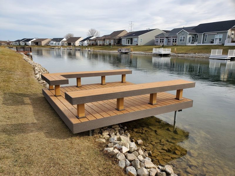 Dock with benches along a canal, houses in background. Brown deck and benches, cloudy sky.
