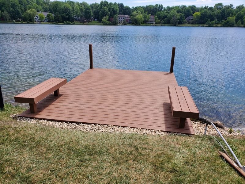 Brown dock with benches extends into a lake, surrounded by green grass and trees.