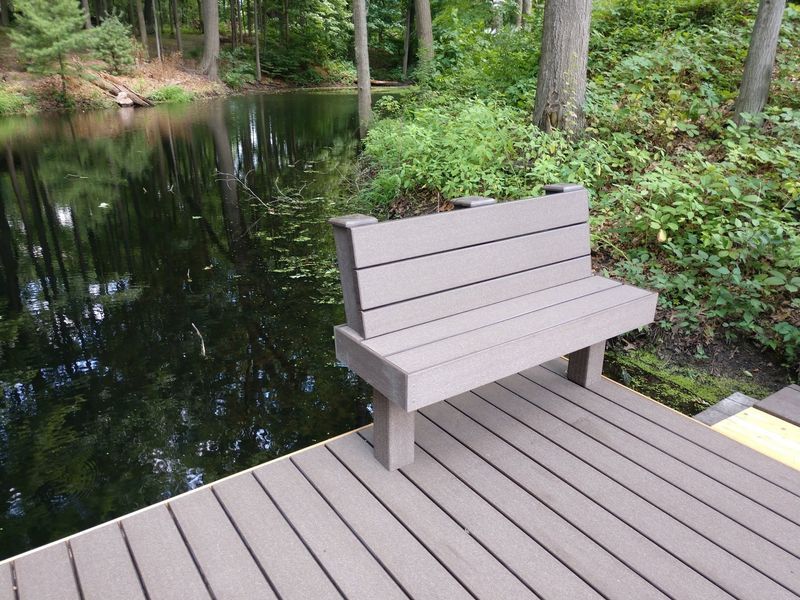 Gray bench on a wooden dock overlooking a still, dark lake surrounded by trees.