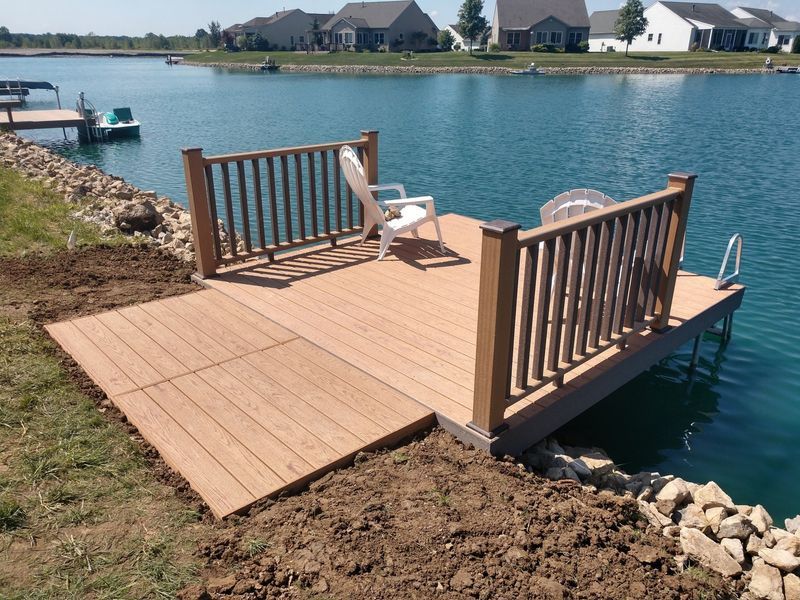 Wooden dock with railings, chair, and a ramp extending to the lawn, at the edge of a blue lake.