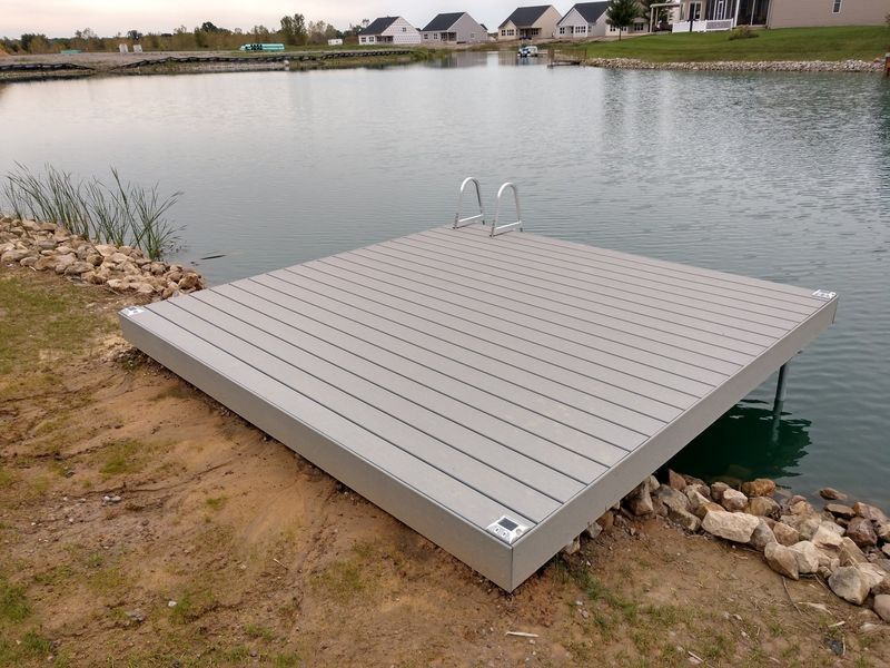 Square dock with ladder extends into calm water. Gray composite decking, low-lying, with residential homes in the background.