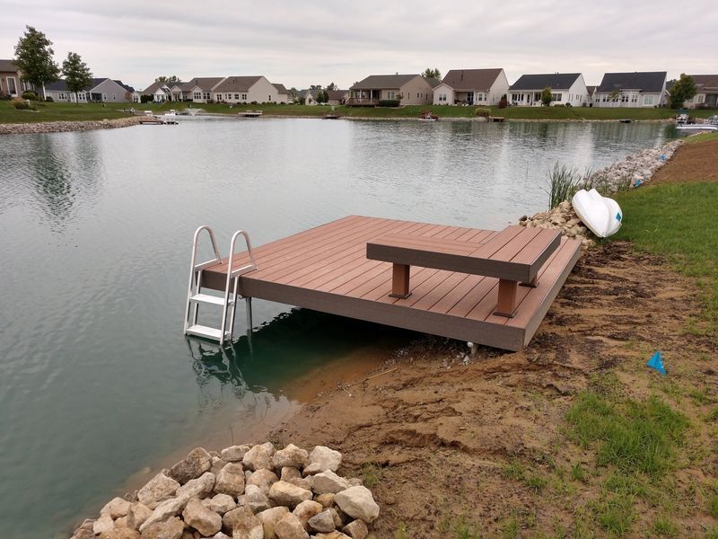 Dock with bench, ladder, and small boat on a lake with houses in the background. Brown, gray, and blue.