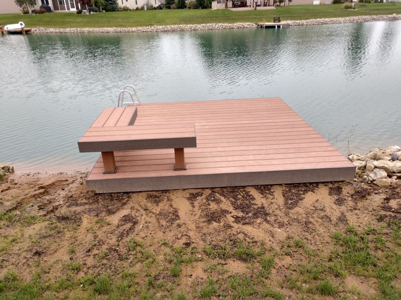 Wooden dock with built-in bench on a lake. Brown ground in front, water in background.