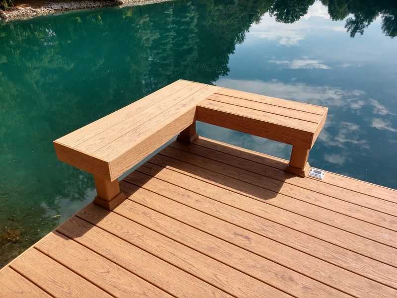 Wooden dock with built-in bench overlooking calm, turquoise water. Reflective sky visible in water.