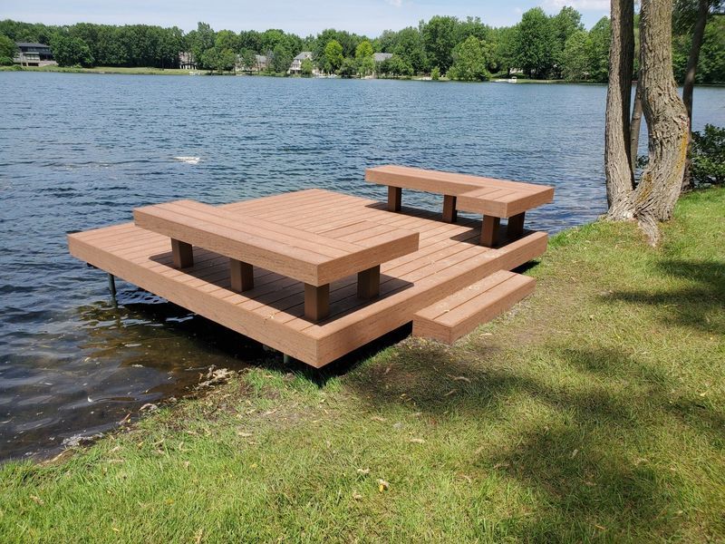 A brown dock with attached picnic table and benches over a lake, green grass in foreground.