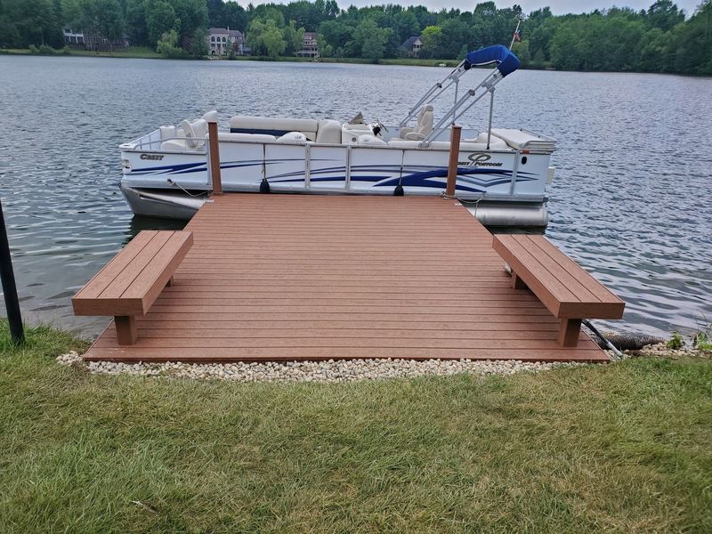 Wooden dock with benches and a pontoon boat on a lake. Brown deck, green grass, and blue water.