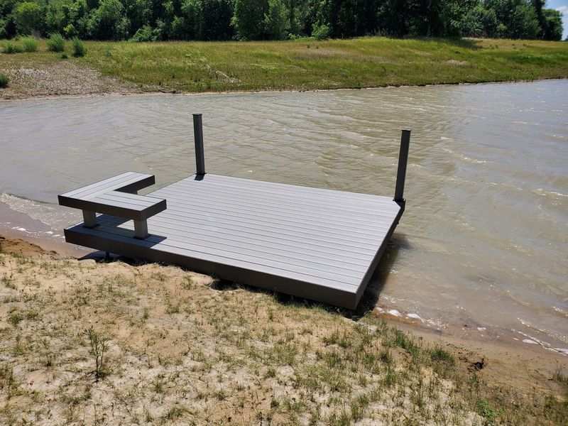 A gray dock with a bench sits at the edge of a muddy body of water, grassy bank in background.