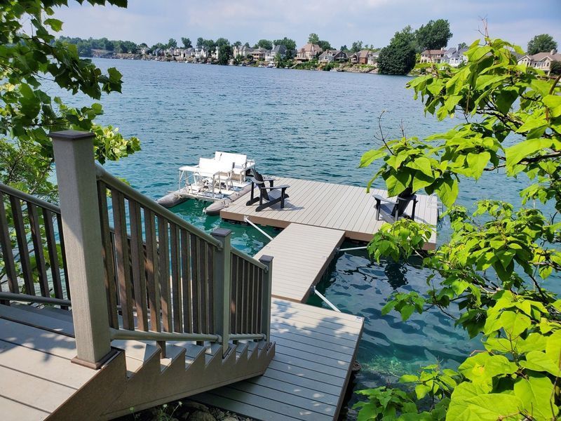 Wooden dock with stairs, chairs, and lake view. Houses visible in the background. Sunny day.