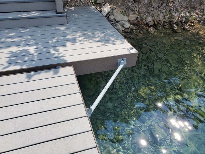 Gray composite dock with support arm over clear water, rocky shoreline in background.