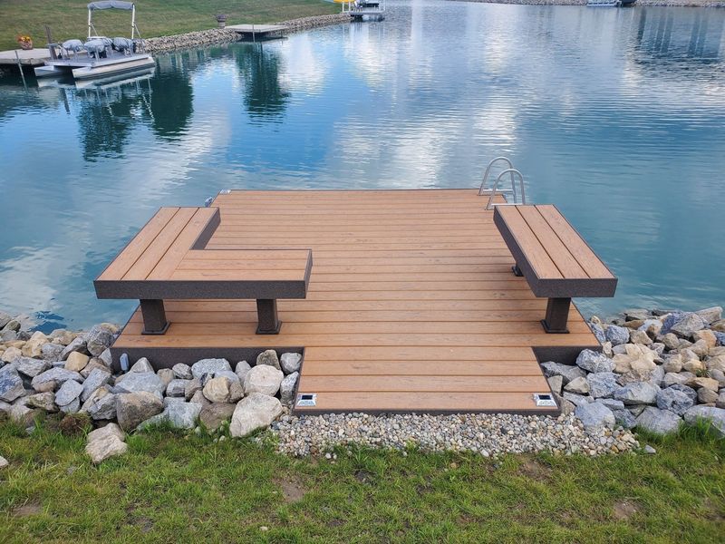 Wooden dock with two benches, set on a lake. Rocks line the shore, with a boat and fountain in the background.