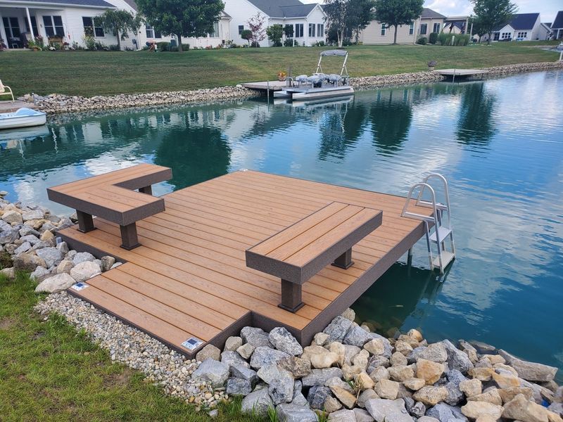 A brown composite dock with seating extends into a body of water.  A boat and houses are in the background.