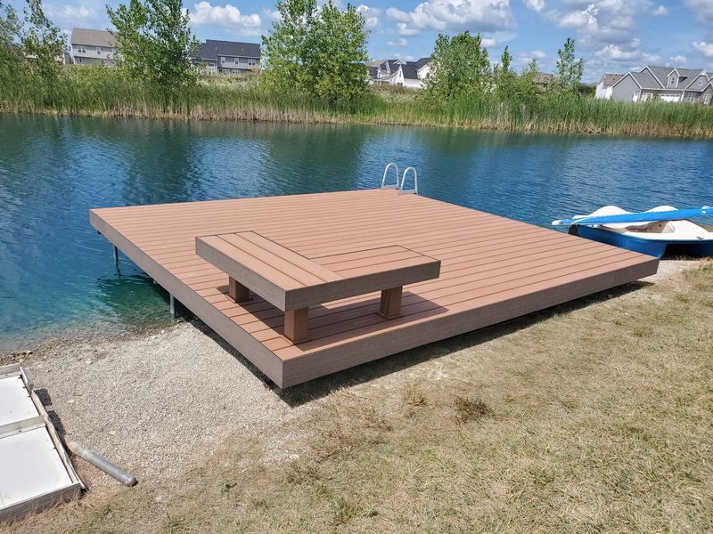 Dock on blue water with a built-in table. Reeds, trees, and houses in the background. Sunny day.