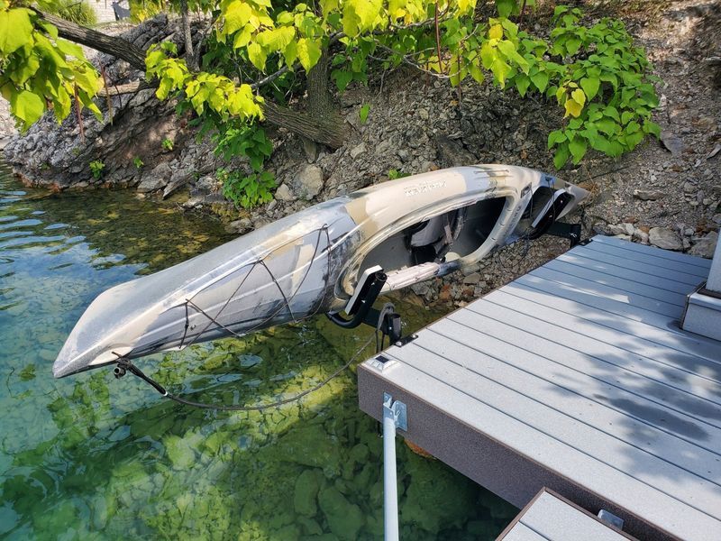 Kayak on a dock near water, camouflaged paint, supports extended. Green and brown vegetation in the background.
