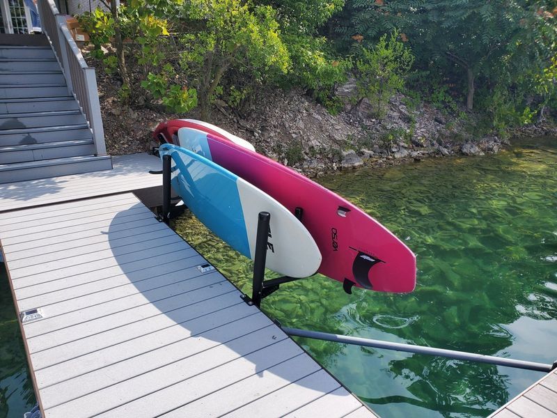 Two paddle boards, pink and blue, on a black rack, dockside, near a staircase and water.