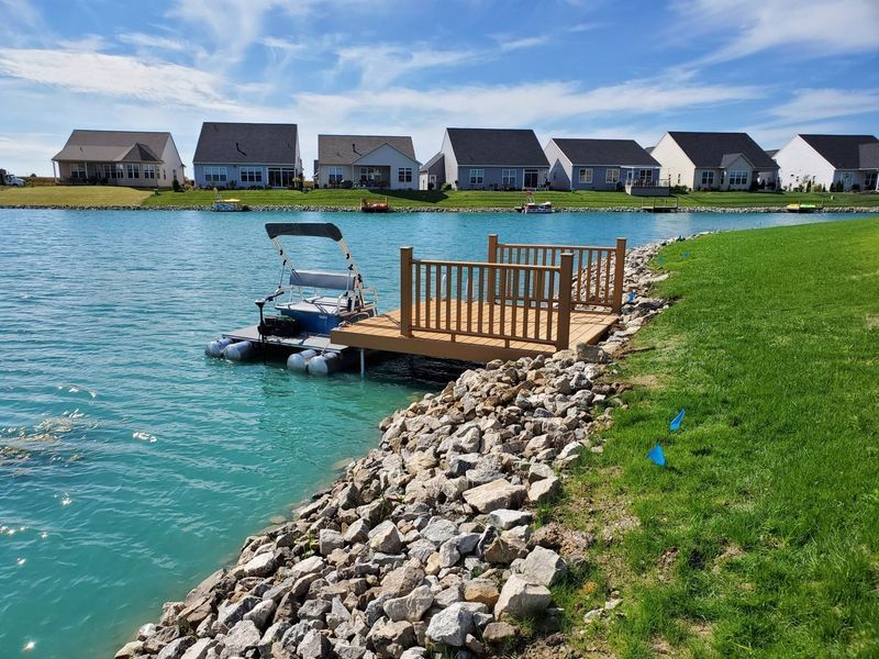 Dock on blue water, rocky shoreline, houses in background.