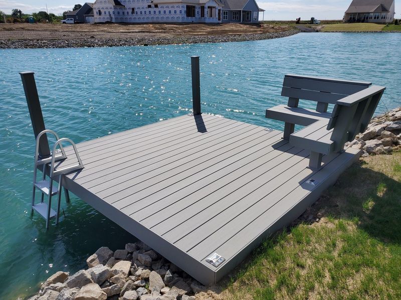 A gray dock with bench and ladder on a turquoise lake. A house is in the background.
