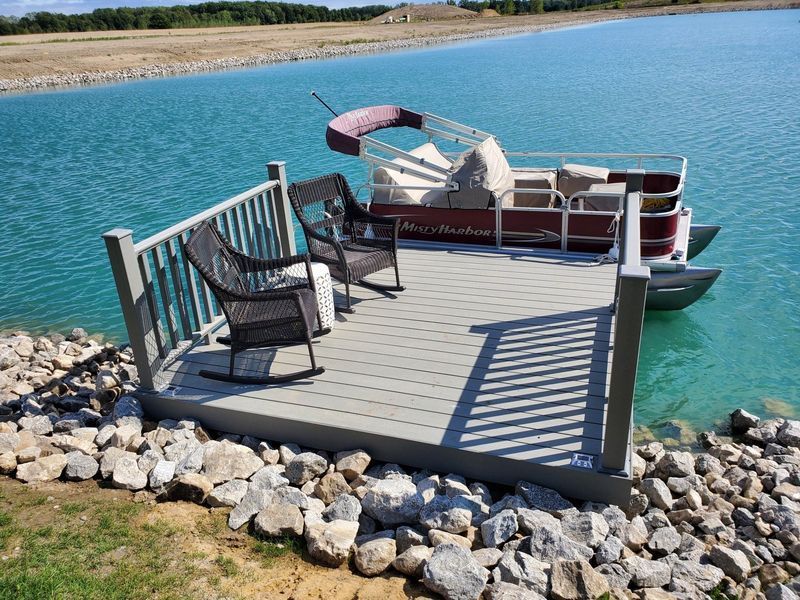 Pontoon boat docked at a small deck with chairs on a blue lake.