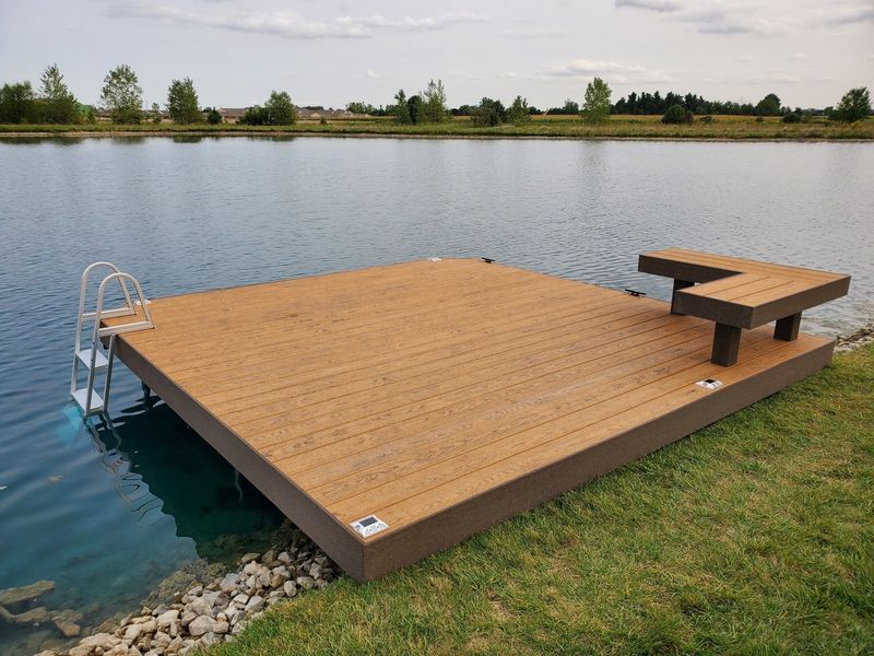 Wooden dock on a lake with a bench and ladder, brown decking, green grass, and blue water.