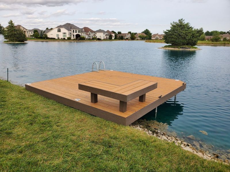 Brown dock with a bench on a lake, residential area in the background.