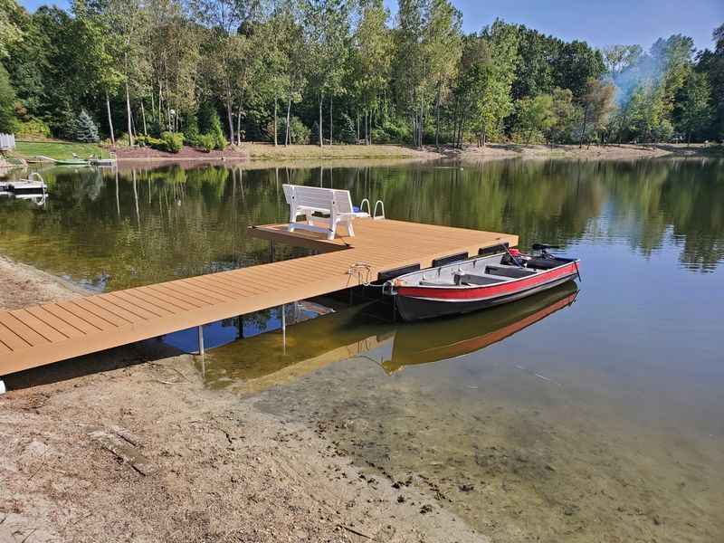 Brown wooden dock with a white bench and small motorboat on a lake, surrounded by trees.