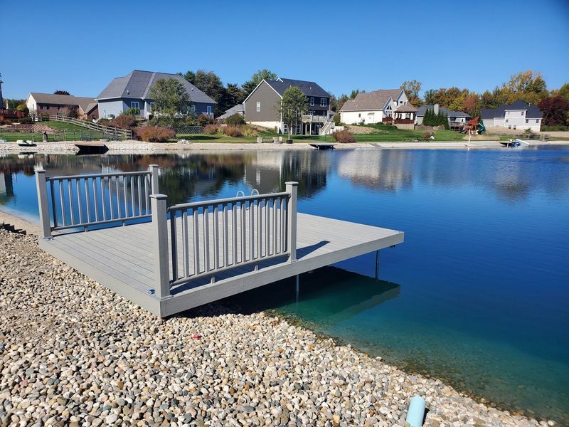 A dock with gray railings extends into a blue lake, gravel shoreline, houses in background under blue sky.