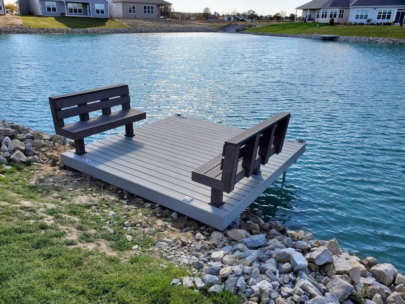 Dock with two benches over a lake. Gray deck with dark brown benches. Rocky shoreline and houses in the background.