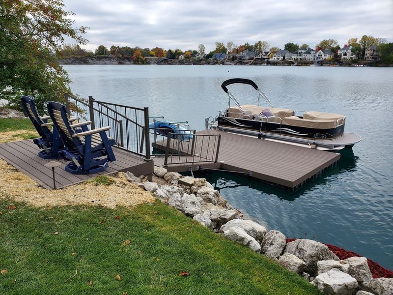 Dock with pontoon boat, lounge chairs, and water access. Houses line the opposite shore. Cloudy day.