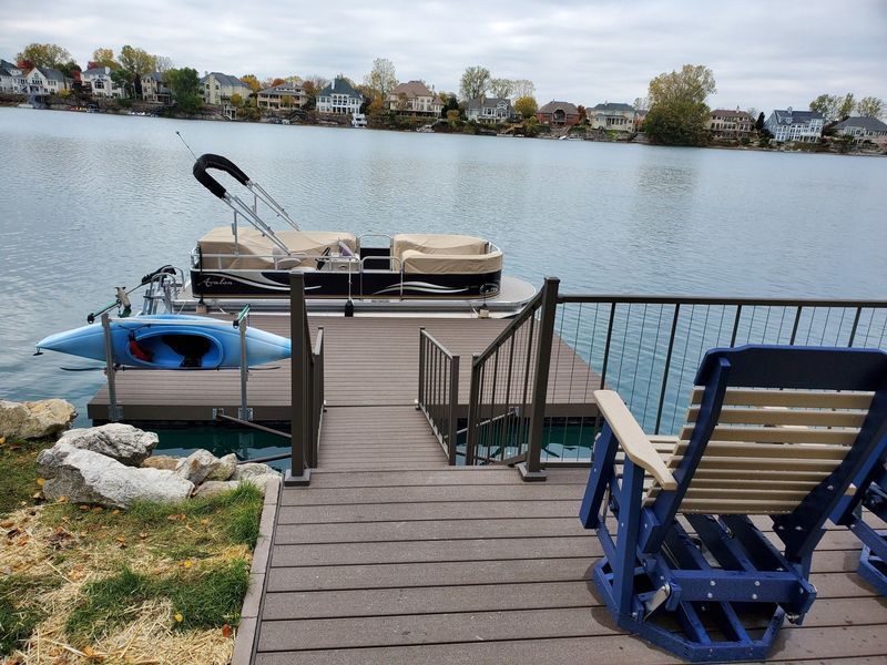 Dock with boat and kayak on water, wooden stairs, and seating overlooking lake with houses in the background.