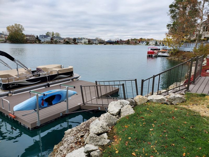 A lakefront dock with a kayak and a pontoon boat. Brown dock, blue water, cloudy sky, houses in background.