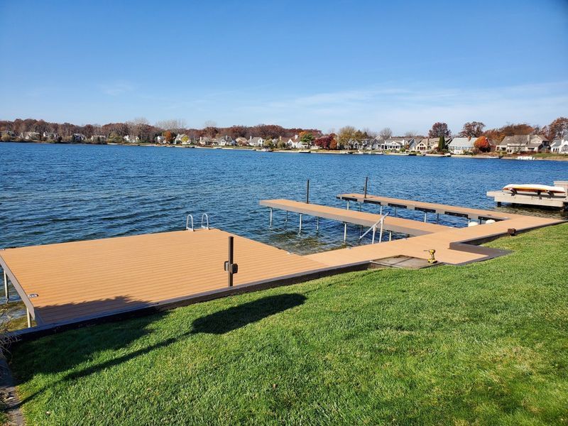 Lakeside dock with brown decking and metal supports, grassy shoreline, blue water and sky, houses in the distance.