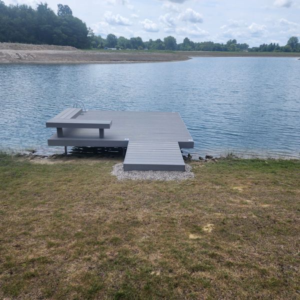 Gray wooden dock with bench on lake edge, grass in foreground, blue water and sky.