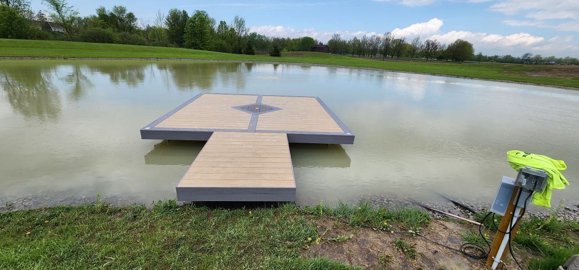 Wooden dock on a calm pond, surrounded by green grass and trees. Blue sky overhead.
