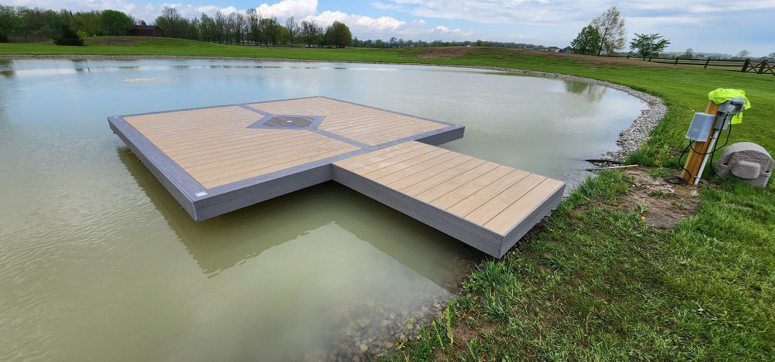 Floating dock on a pond with a grassy bank and trees in the background. Cloudy sky.