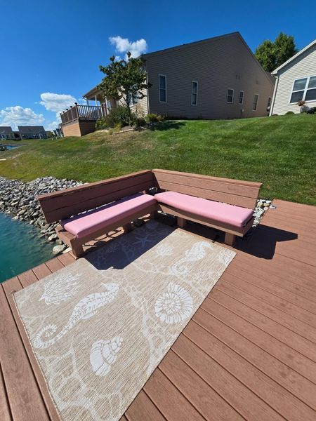 L-shaped wooden bench with pink cushions on a deck overlooking water. Beige rug with sea-themed designs.